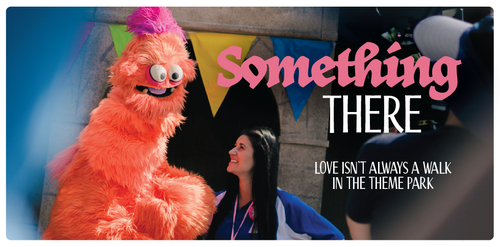 A promotional graphic for a short film features a tall orange monster puppet standing next to an amusement park employee in front of a castle. Text reads “Something There: Love isn’t always a walk in the theme park.”