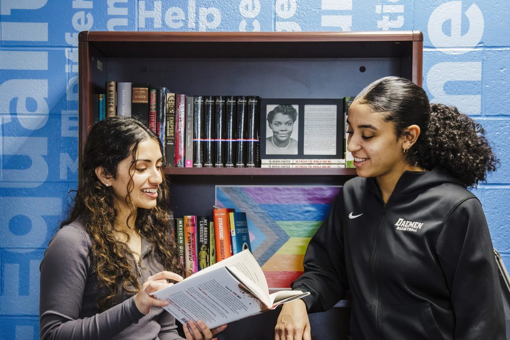 Two students standing in the HOPE Center, engaged in conversation about a book one of the students is holding open