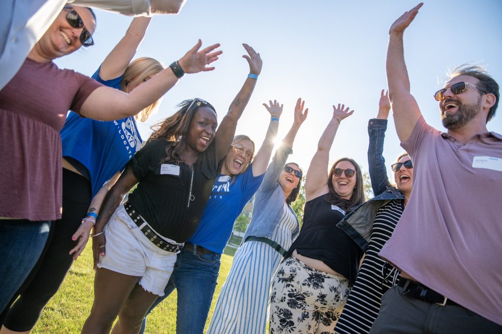 Six alums standing together raising one hand into the air and cheering