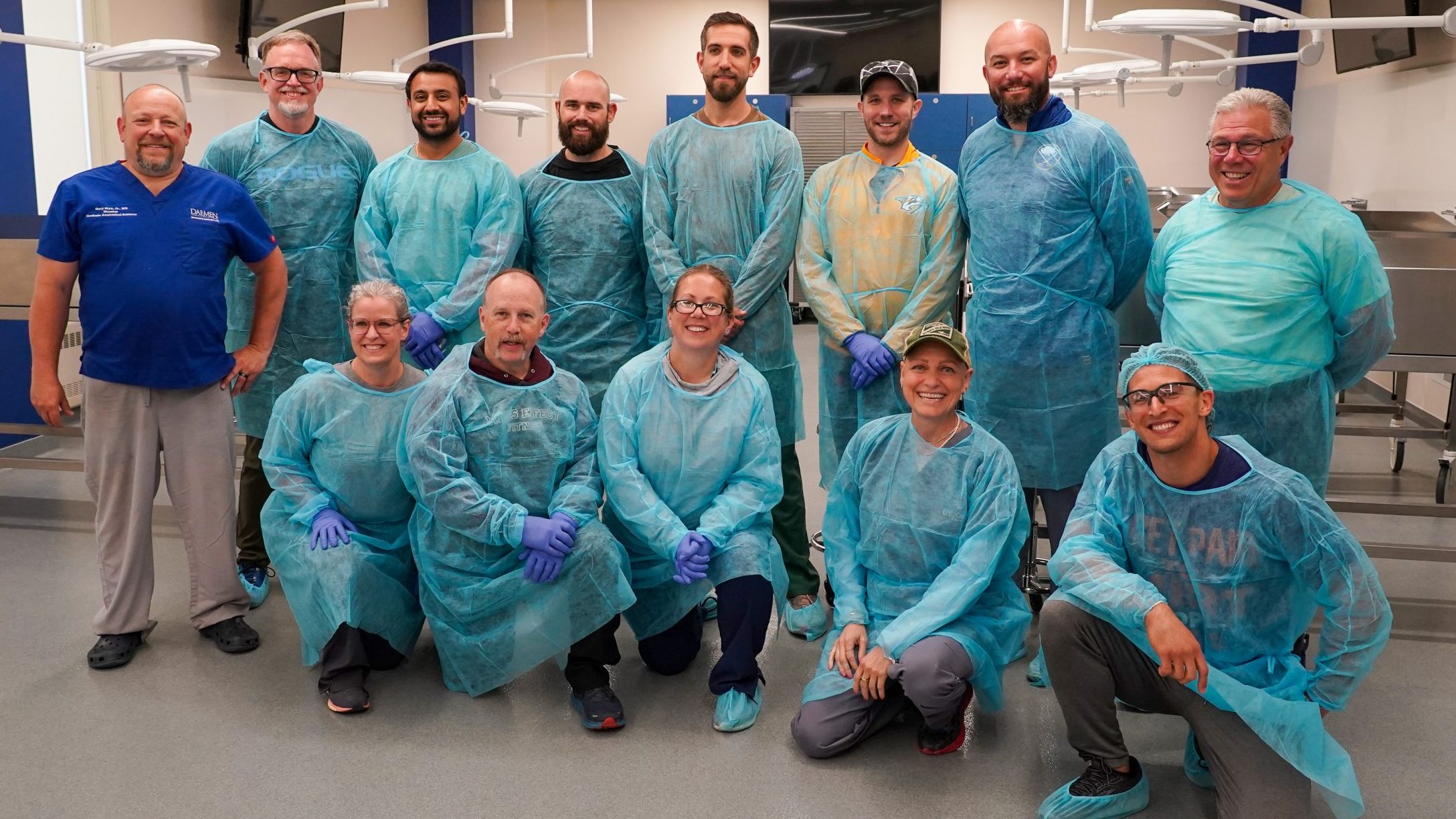 Sue Falsone and Dr. Gary Styn pictured with 11 attendees dressed in blue disposable isolation gowns inside the anatomy lab